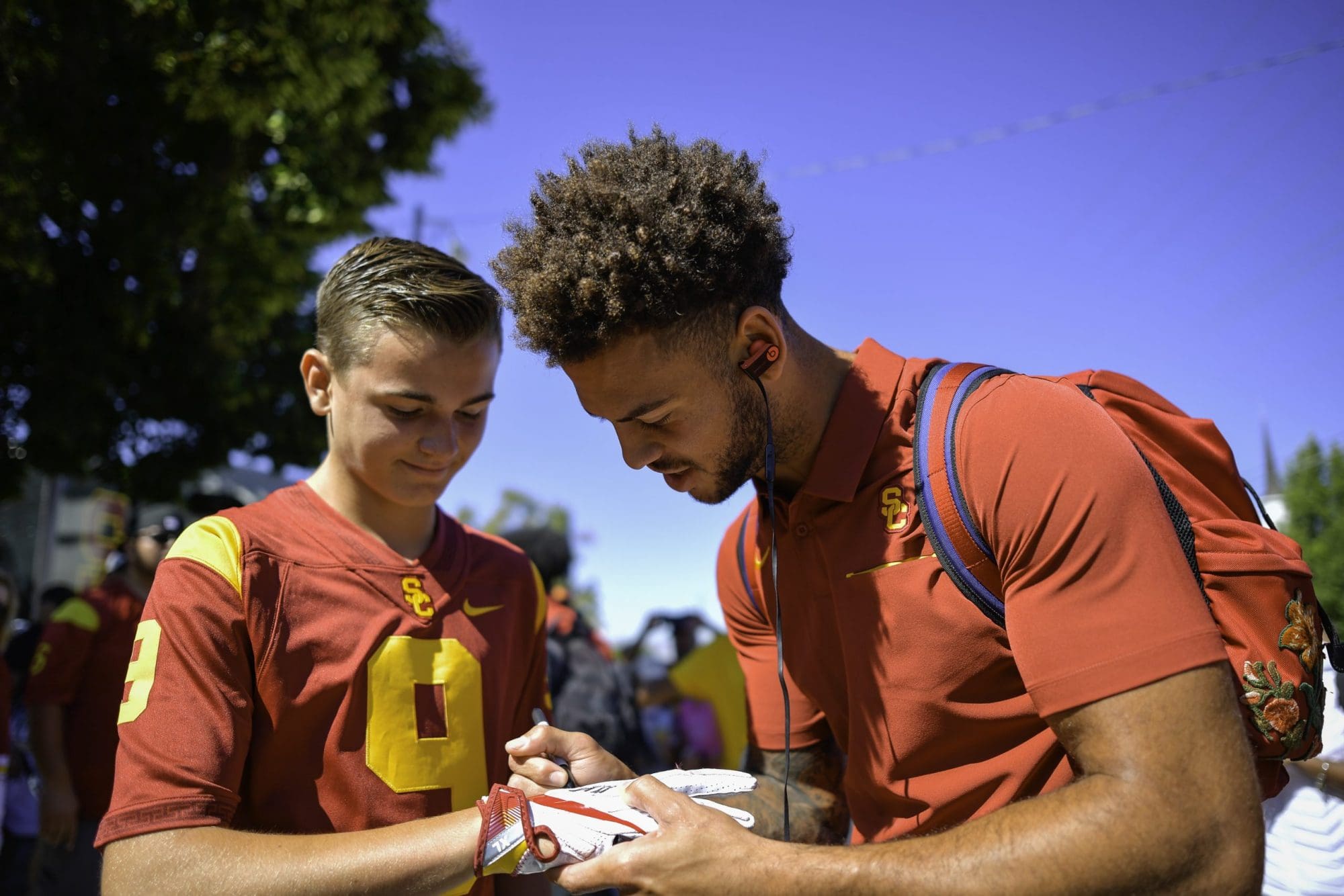 A young male athlete in a red and gold football jersey autographs memorabilia for a fan, also wearing a similar jersey, during a sunny outdoor meet-and-greet event.