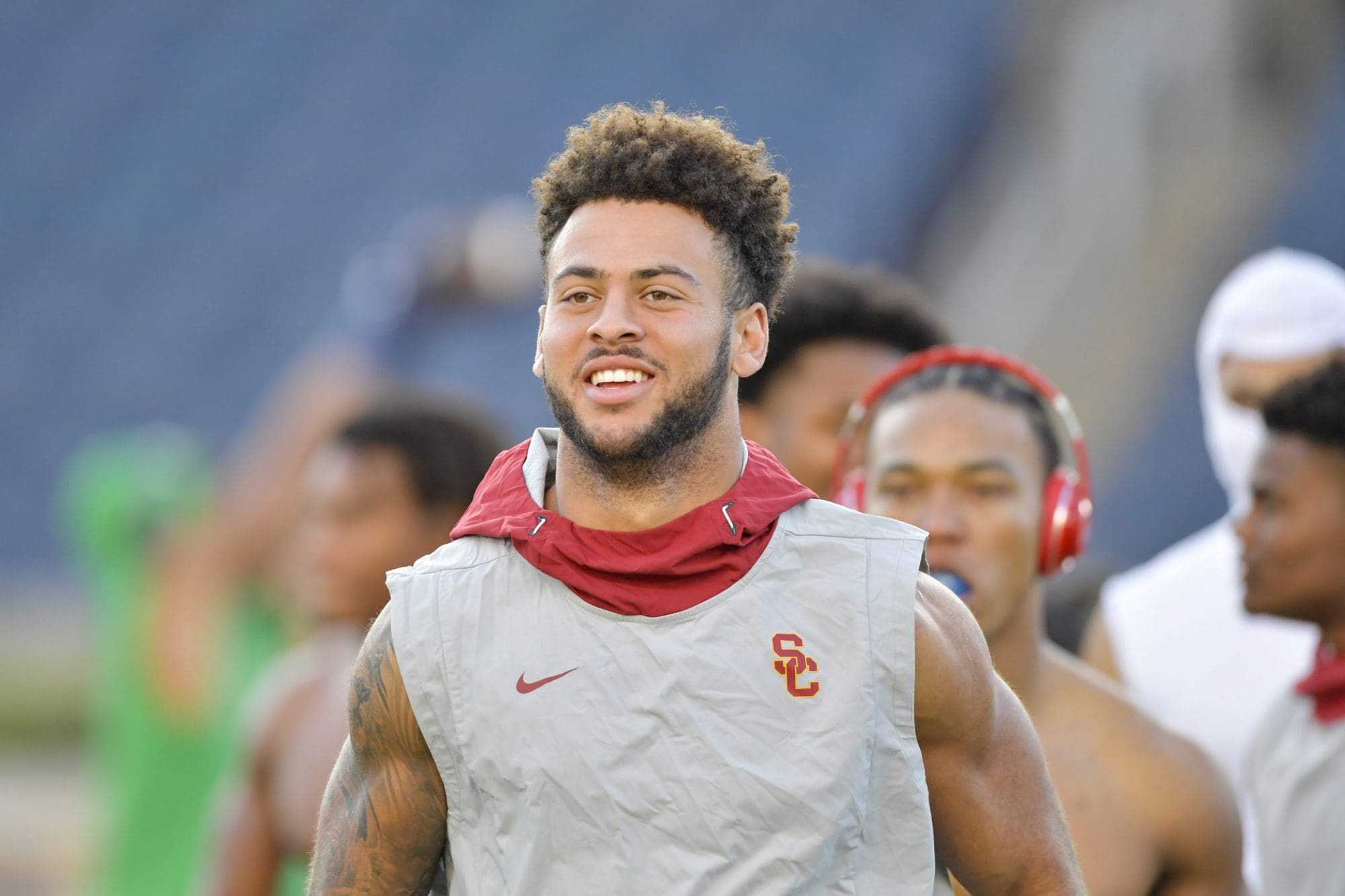 A young athlete with curly hair, wearing a red and gray usc football uniform and a padded vest, smiles while standing on a field with other players in the background.