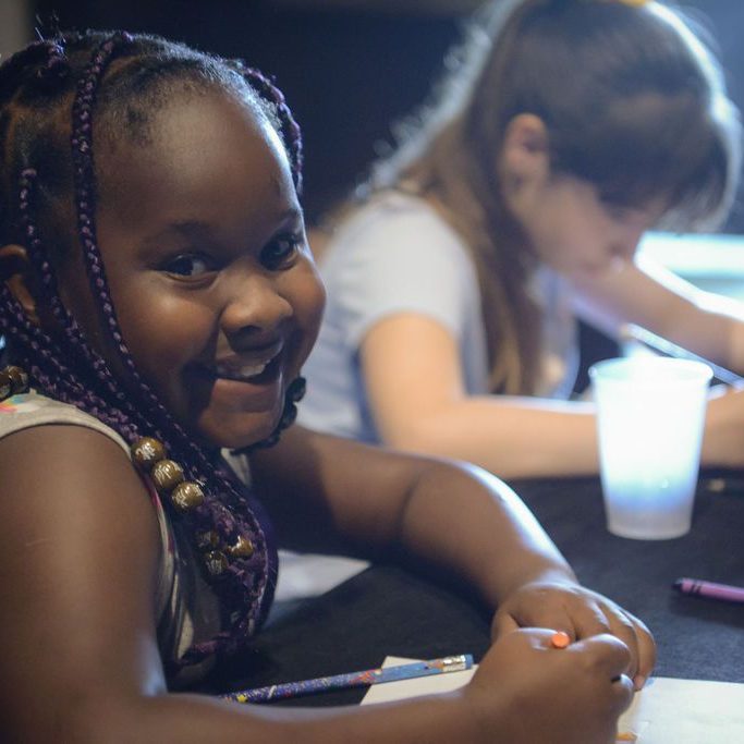 A young girl with braided hair and colorful beads smiles at the camera while drawing with crayons at a table, another girl focused on her drawing in the background.