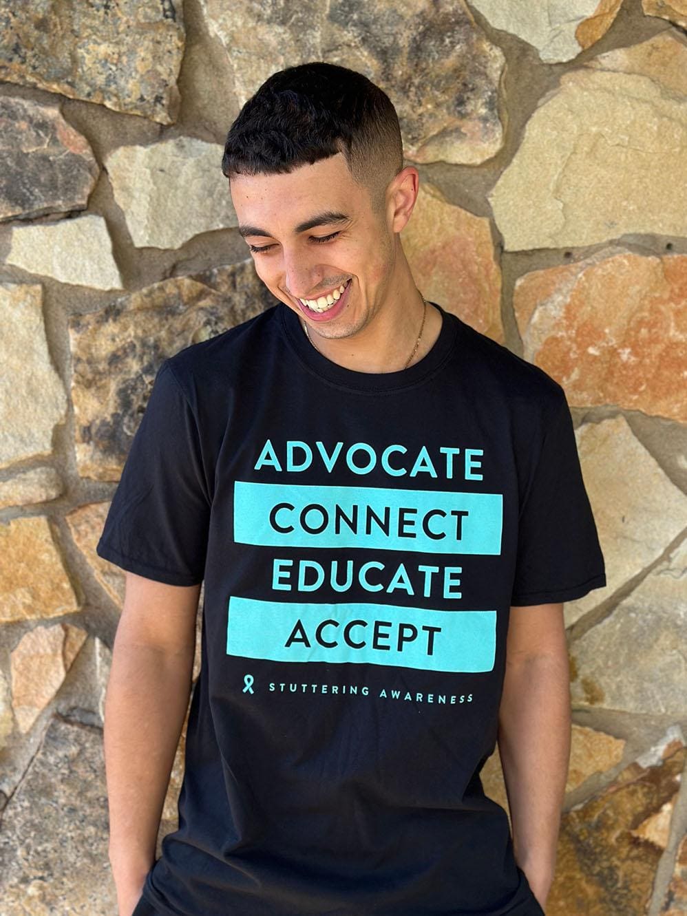 A young man smiling, wearing a black Advocate Tee with "advocate connect educate accept & stuttering awareness" printed in teal and white text, standing against a stone wall background.