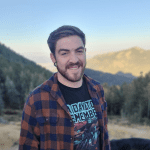 Alden Wheeler, a smiling man with a beard sporting a plaid shirt and a graphic tee, stands outdoors with a forested mountain landscape unfolding behind him during sunset.