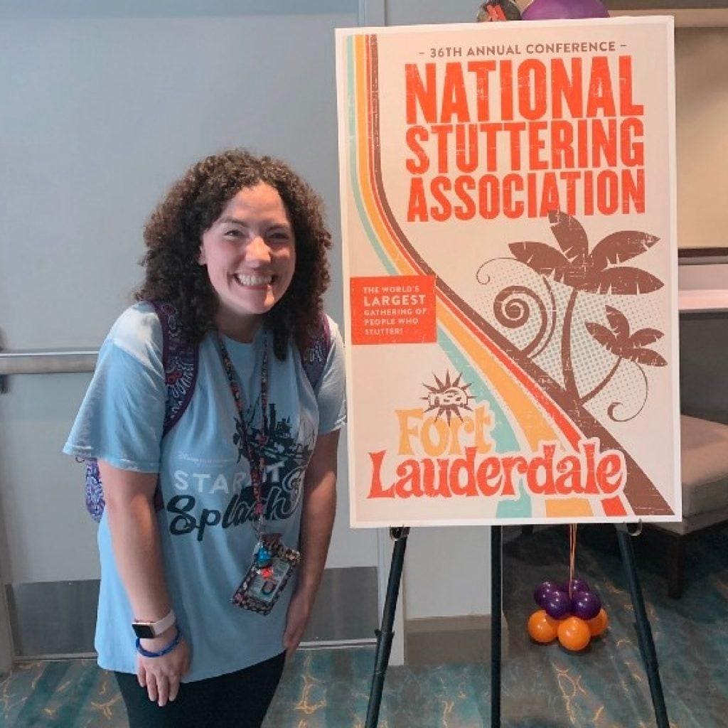 A smiling woman with curly hair, wearing a blue t-shirt and a lanyard, stands next to a poster announcing the "36th annual conference national stuttering association" in fort lauderdale.