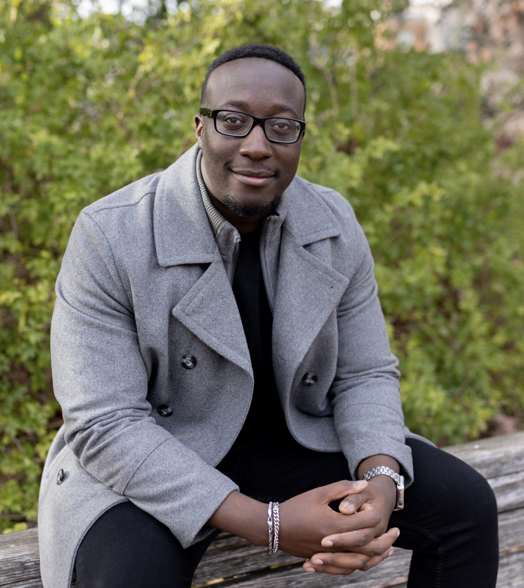 A person with short hair, glasses, and a friendly smile sits on a wooden bench outdoors. They are wearing a gray coat over a black shirt and have their hands resting in their lap. Green leafy bushes are in the background.