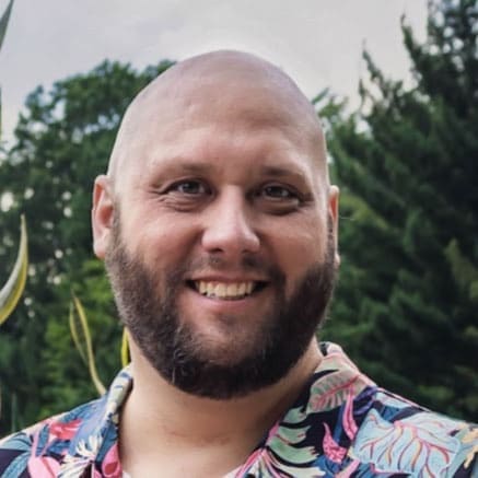 A smiling man with a bald head and a beard stands outdoors. He is wearing a colorful, floral-patterned shirt, and trees are visible in the background. The atmosphere is bright and cheerful.