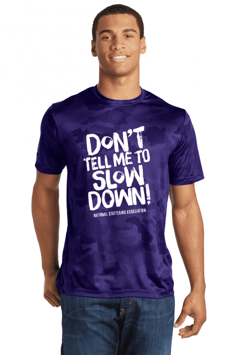 A young man wearing a purple Don't Tell Me to 'Slow Down' Sport-Tek Tee with the text "don't tell me to slow down!" along with "national stuttering association" printed on it, smiling at the camera.