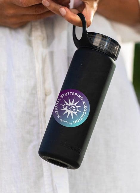 Person holding a black water bottle with a WE Stutter Stickers logo, partially concealed by their white shirt. visible against a blurred green background.