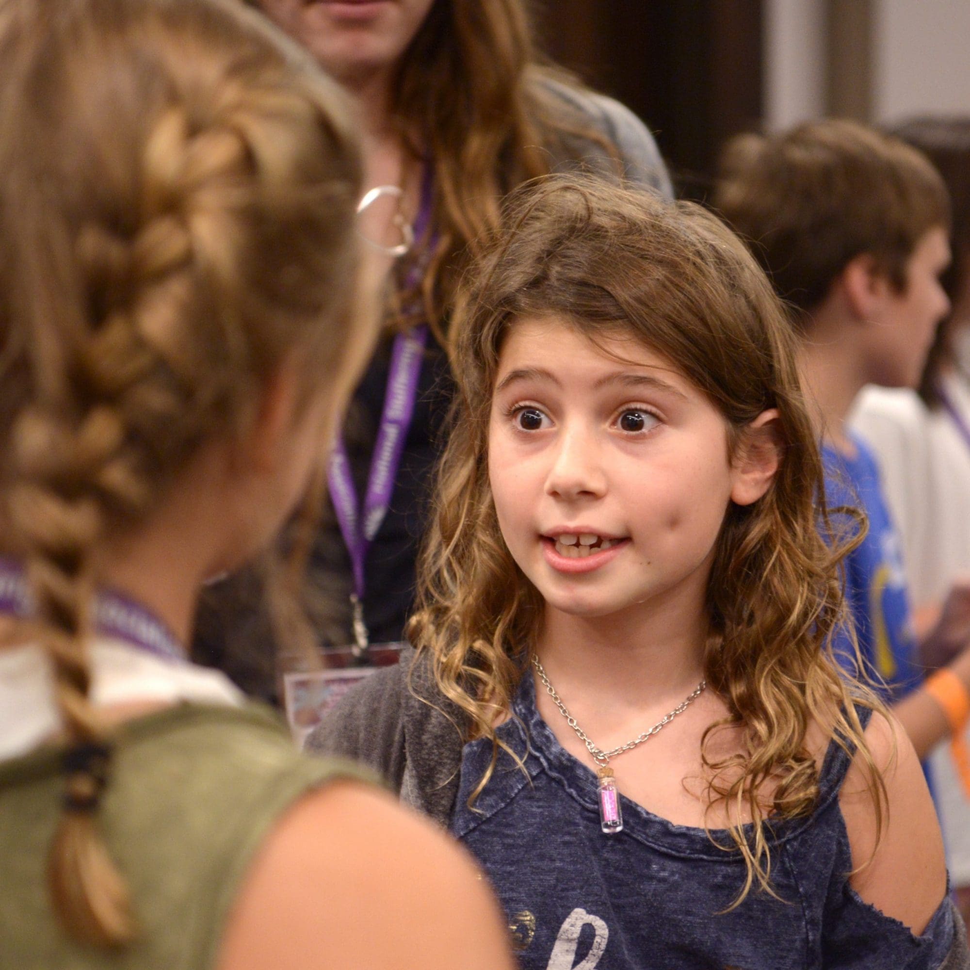 A young girl with curly brown hair and a blue shirt speaks animatedly to another child, whose back is to the camera. They appear to be engaged in a lively conversation in a crowded room, promoting