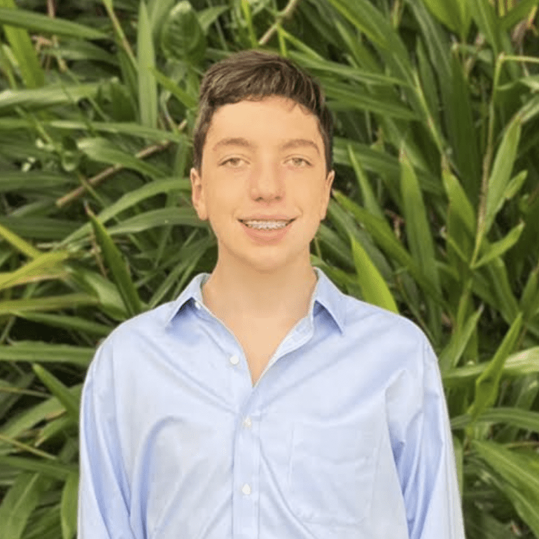 A young man smiles gently at the camera, wearing a light blue shirt for his "Meet the Team" photo. He is positioned in front of a lush, green leafy background, enhancing the natural