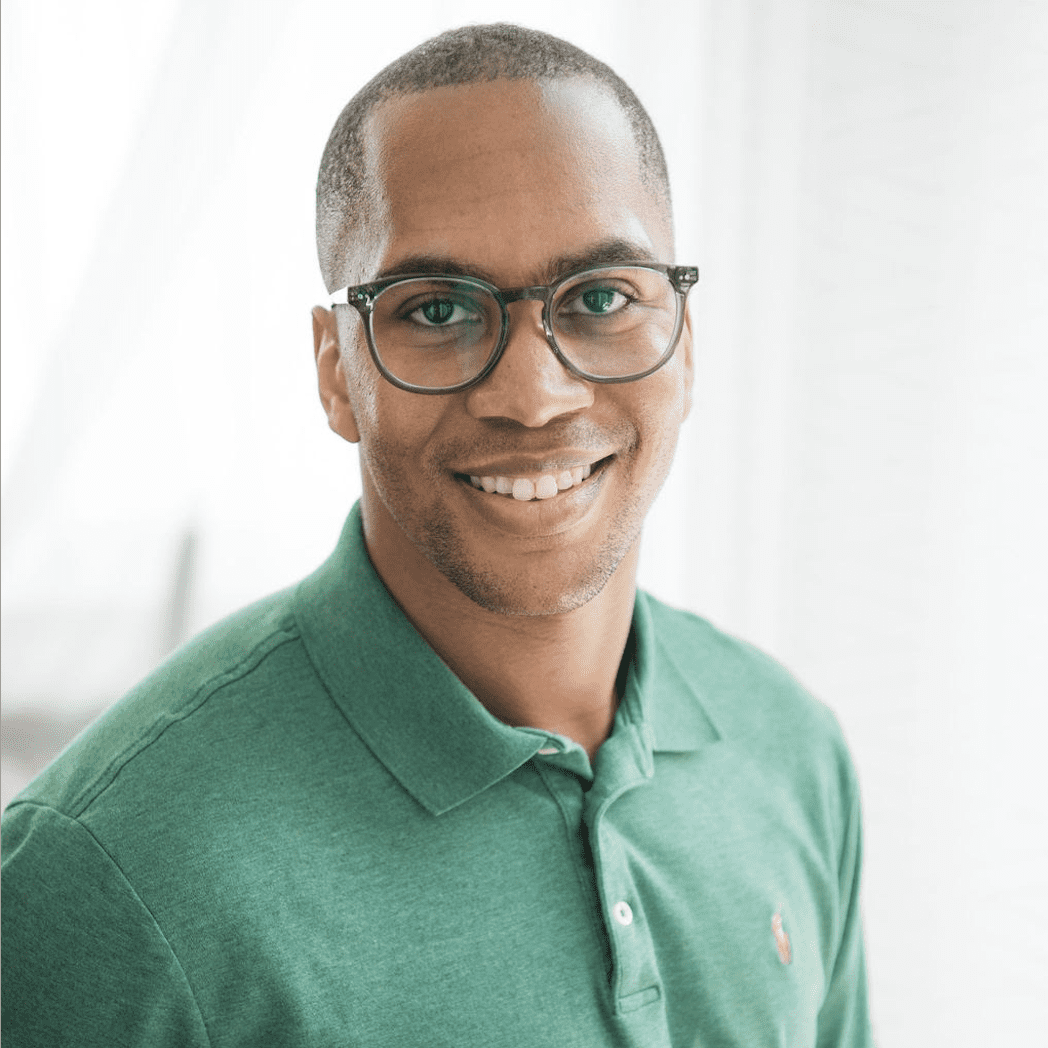 A smiling black man wearing glasses and a green polo shirt, with a softly blurred white background. he appears friendly and approachable.