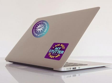 A closed laptop with a gold finish, decorated with two WE Stutter Stickers, one from the national stuttering association, and another stating "i am not my stutter, not me.