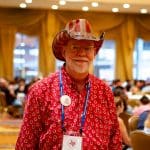 Russ, a man with a white beard and glasses, stands in a room with other people, smiling at the camera. He wears a red shirt with a white horseshoe pattern, a lanyard with a badge, and a red and white striped cowboy hat. The background is blurred with warm lighting and curtains.