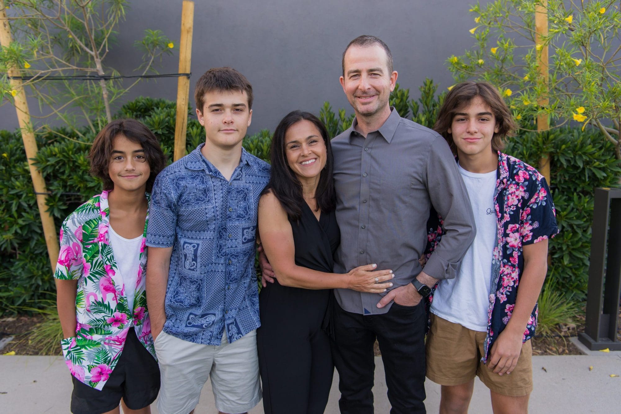 A happy family of five, with three teenage boys and their parents, standing together outdoors, joyfully smiling at the camera in front of a garden and a dark wall.