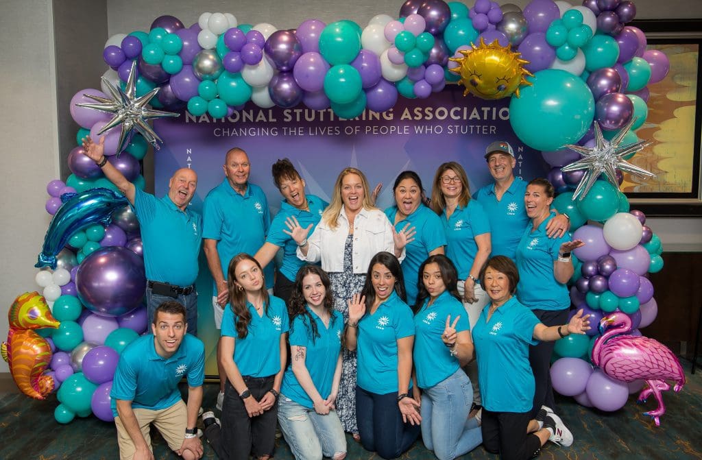 Group of cheerful people in blue and turquoise shirts, gathered around a backdrop with balloons and a banner saying "Meet the Team - National Stuttering Association: Changing the Lives of People Who Stutter".