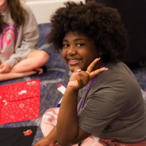 A young teen girl with curly hair sitting cross-legged on the floor, giving a peace sign and smiling at the camera in a room with other sitting participants.