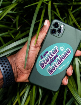 A hand holding a smartphone with a WE Stutter Stickers case that reads "if you stutter you are not alone" over a background of green leaves. the phone case has a url: yesstutter.org.
