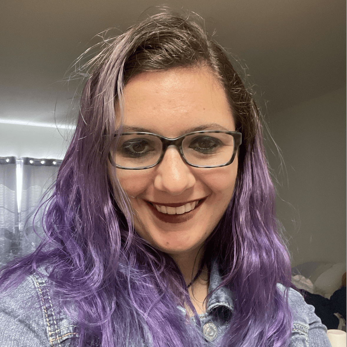 Elizabeth Minton, a smiling woman with purple hair and glasses, wearing a denim jacket, is indoors with soft lighting and a blurred background.