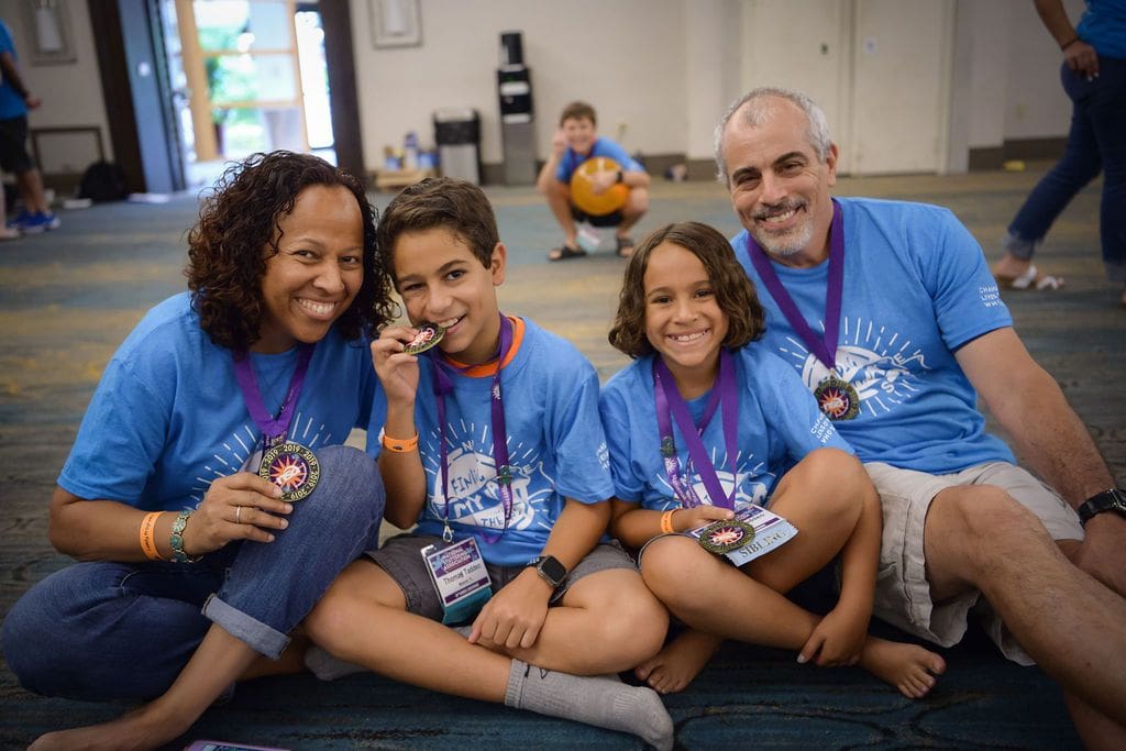 A joyful family of four sitting indoors, wearing matching blue t-shirts and medals, smiling at the camera. Two adults flank a young teen and a child, all proud to donate to the NSA.