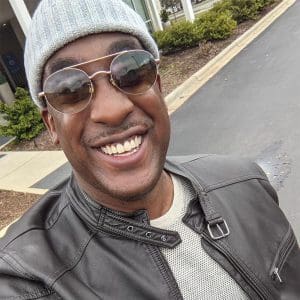 A smiling man celebrating career success, wearing sunglasses, a beanie, and a leather jacket takes a selfie outdoors on a cloudy day.