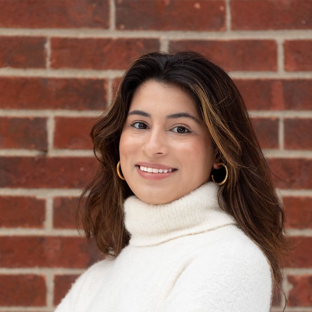 Hispanic woman, with long brown hair, smiling in a white sweater, gold earrings in front of a brick wall.