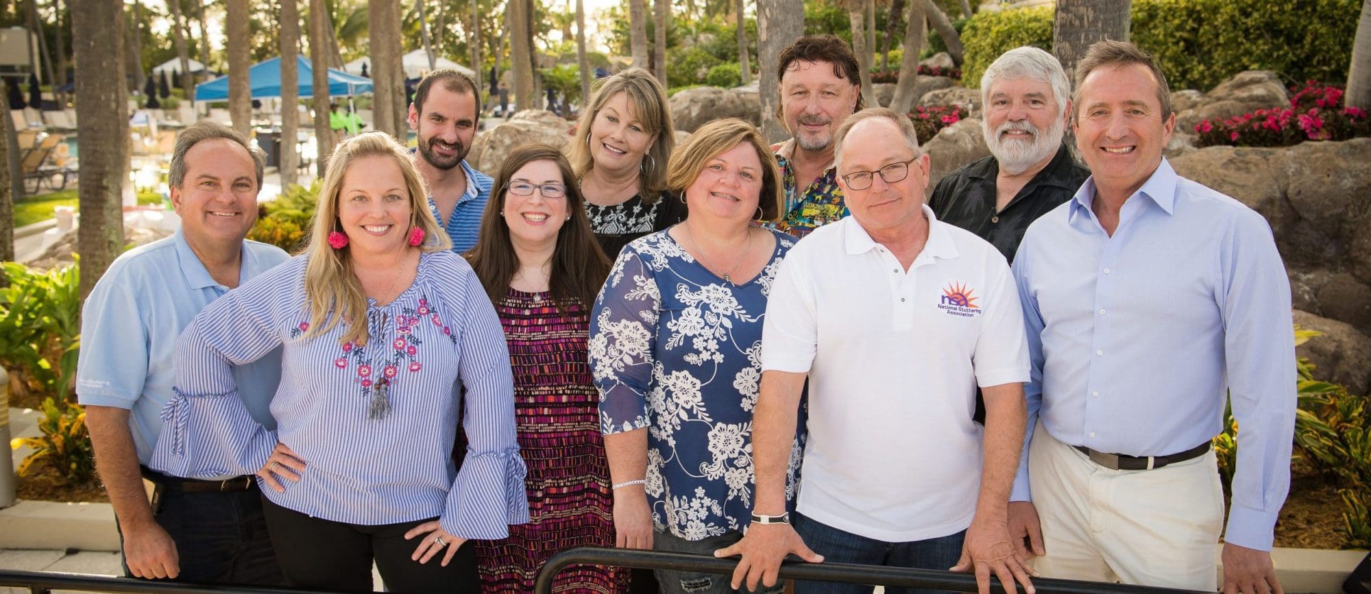 Group of ten adults smiling and posing for a photo outdoors in a sunny garden setting with palm trees in the background.