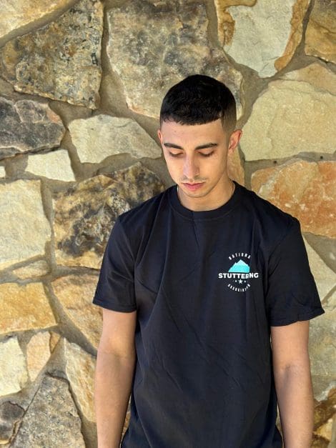 A young man in a black Iceberg Tee standing in front of a textured stone wall, looking down thoughtfully.