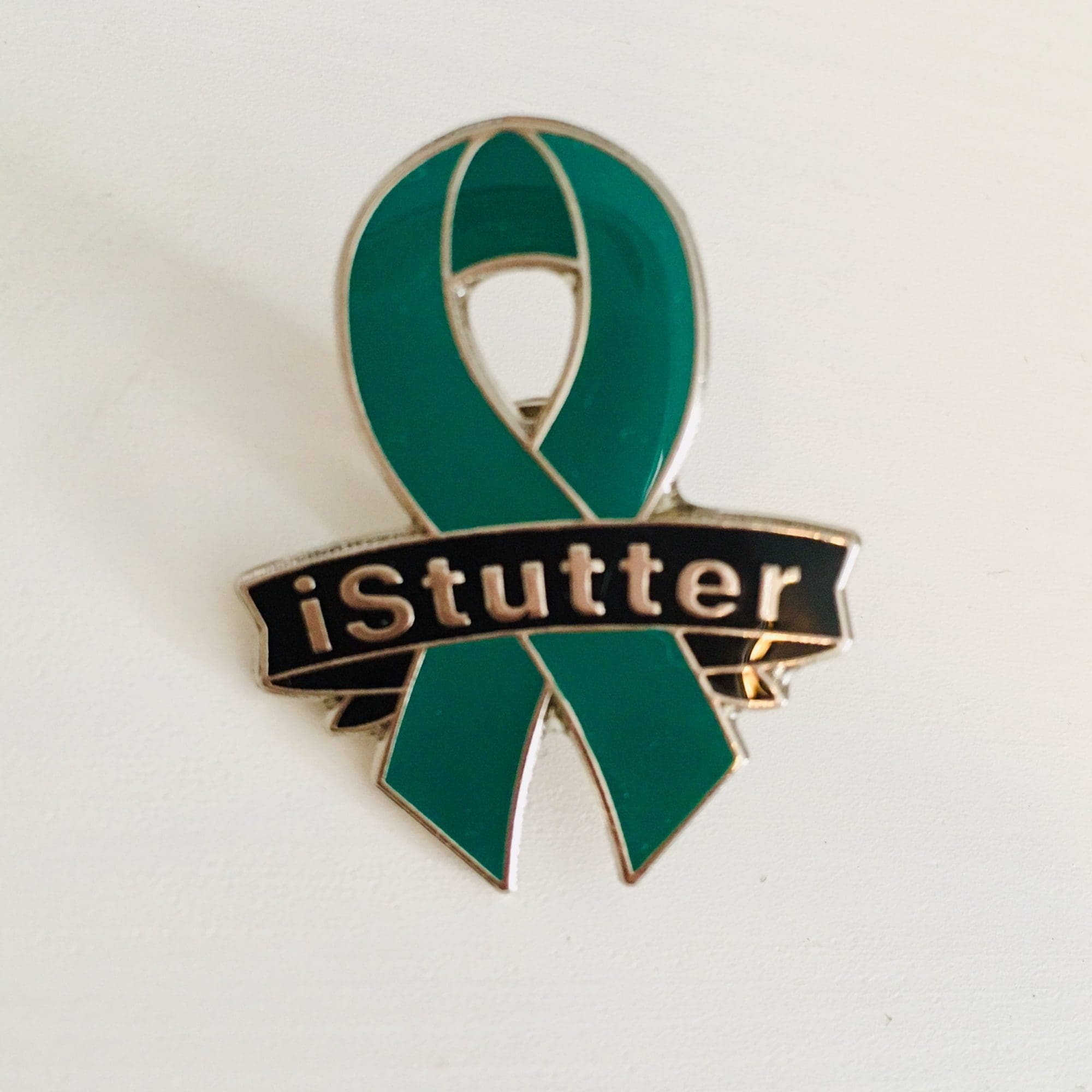 A green awareness ribbon pin with a banner across the center bearing the inscription "If You Stutter, You Are Not Alone" on a white background.