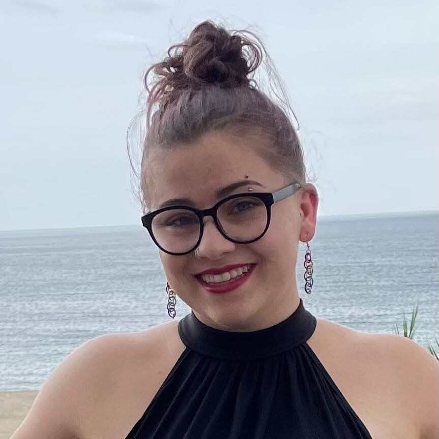 A smiling young woman with glasses, a topknot hairstyle, and chandelier earrings, dressed in a black halter top, posing in front of a calm sea and overcast sky.