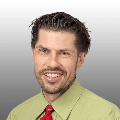 A smiling man with short, neatly styled brown hair, a trimmed beard, and mustache, wearing a light green shirt with a red tie. The background is a simple gray gradient.