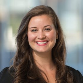 A professional headshot of a smiling woman with long brown hair, wearing a black blouse at the Amplify Conference Portland 2023. She has a friendly expression, standing in front of a blurred city