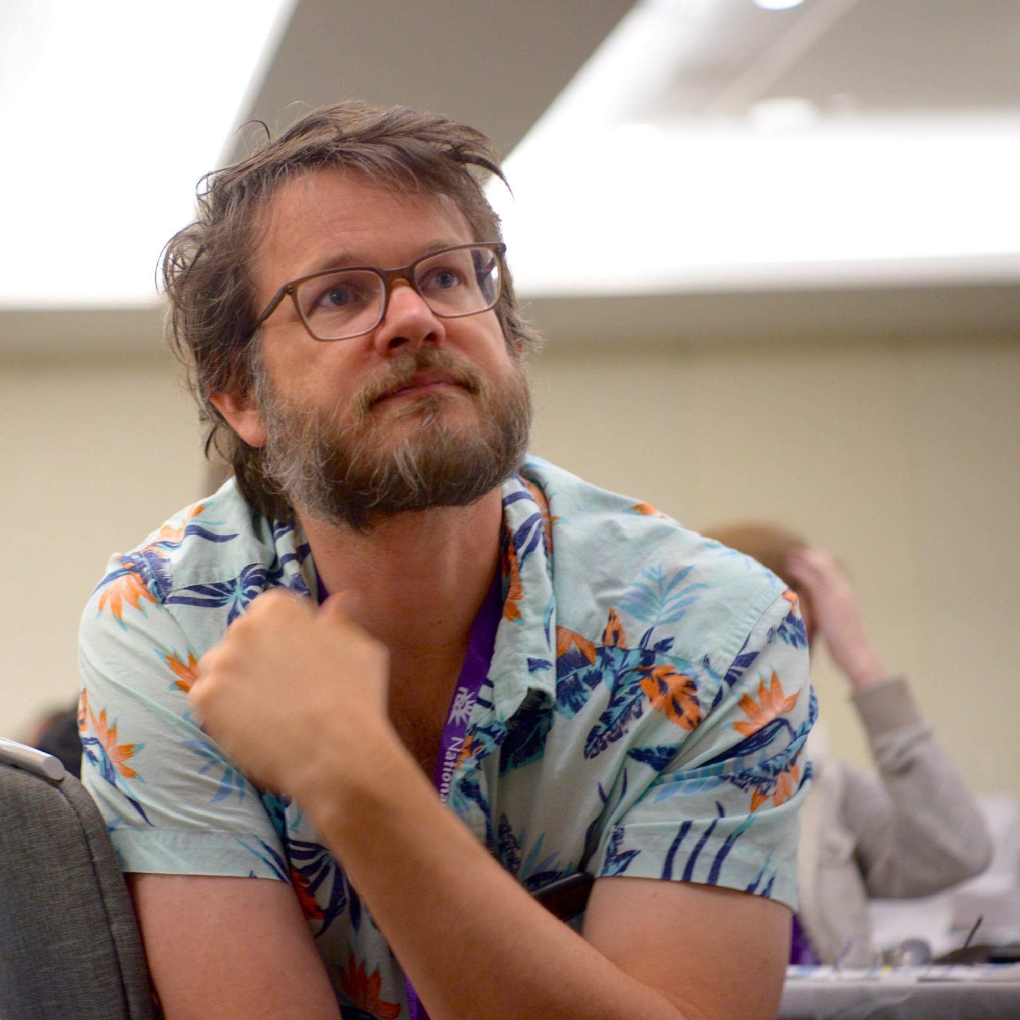 A man wearing a tropical print shirt and glasses looks thoughtfully upwards while sitting in a conference room, pondering his decision to donate to the NSA.