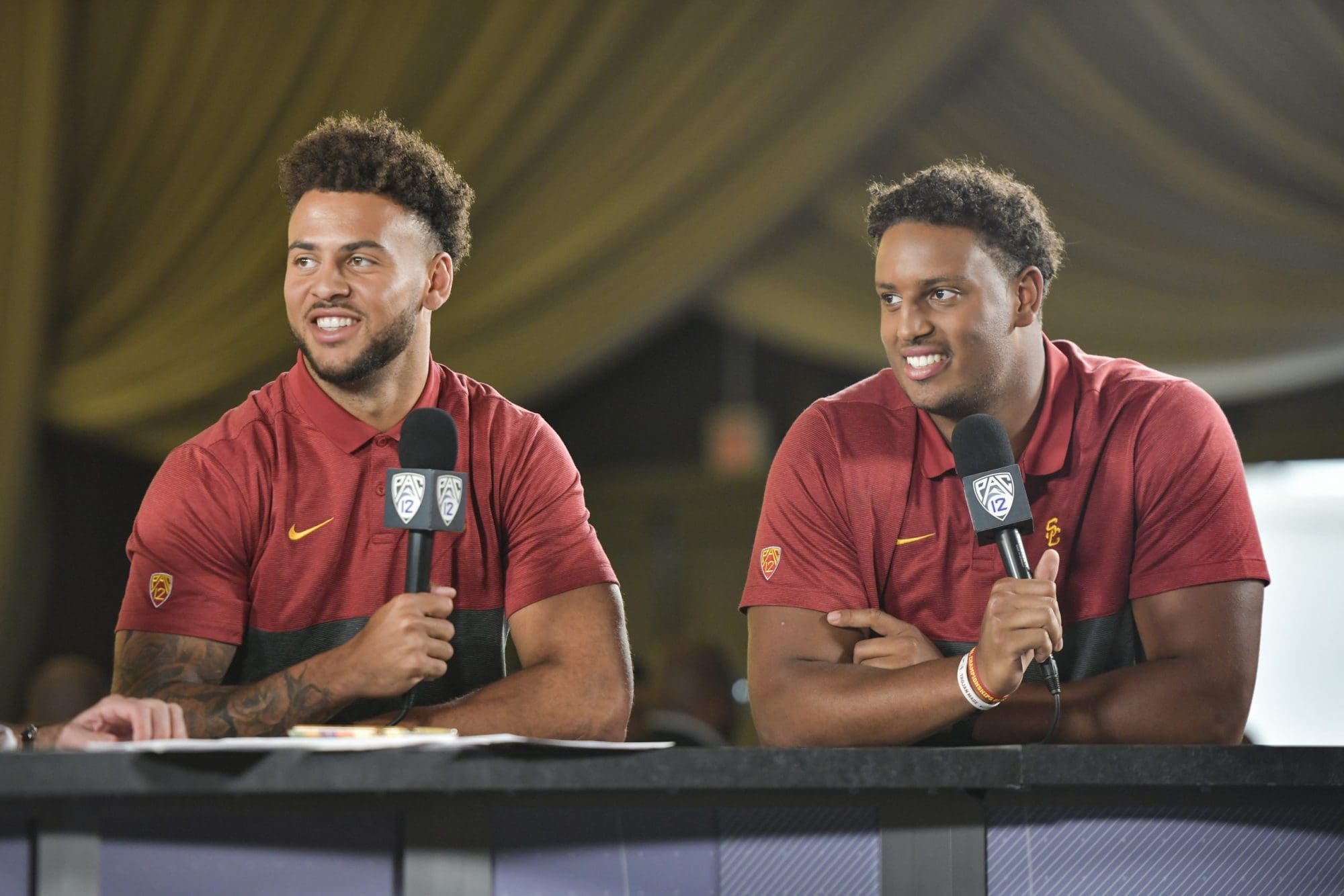 Two smiling athletes in red sports jerseys sit at a table holding microphones, engaged in a discussion during a media event under a large tent.