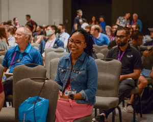 A diverse audience at a conference, with a focus on a smiling woman in the foreground wearing glasses, a denim jacket, and holding a camera. NSA attendees seated around her are listening attentively.