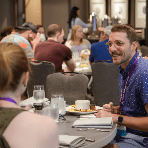 A man in a polka dot shirt smiling at a woman across an NSA conference dining table, surrounded by other attendees engaged in conversation.
