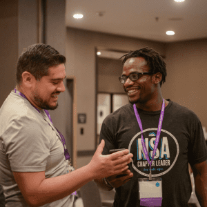 Two men, one wearing a purple lanyard and the other in a black t-shirt with an "NSA chap.r leader" design, are laughing and interacting joyfully at an indoor event.