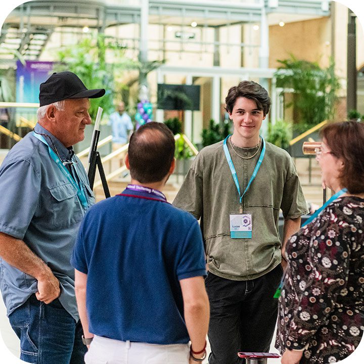 Four people, wearing conference badges, are standing and engaging in a conversation inside a modern building. The background features greenery and glass structures. The group includes three men and one woman, all casually dressed.