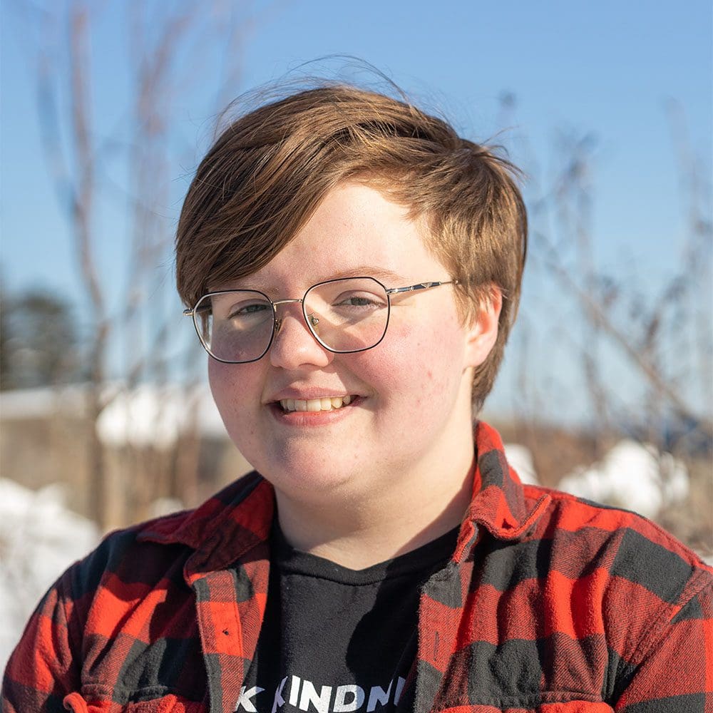 Meet the team: A smiling person with short brown hair and glasses, wearing a red and black flannel shirt over a black t-shirt with the word "kind" visible. The background shows a snowy