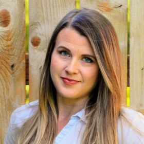 A woman with medium-length brown hair and blue eyes, smiling and wearing a light blue shirt branded with "Amplify Conference Portland 2023," standing in front of a wooden fence.