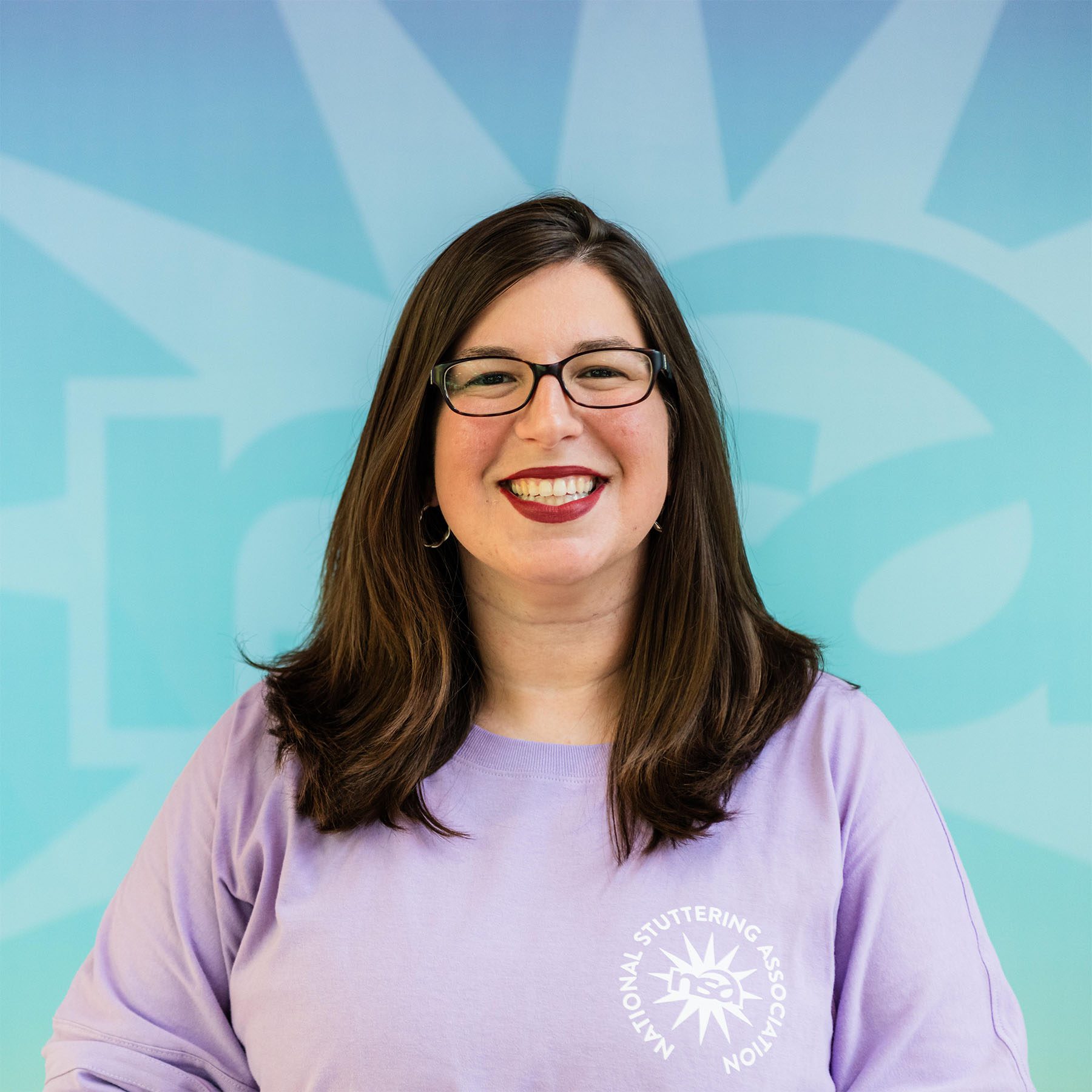 Sarah Onofri, a person with shoulder-length brown hair, is smiling at the camera. She is wearing glasses and a light purple shirt with a logo and text that reads "National Stuttering Association." The background features a teal and light blue design.