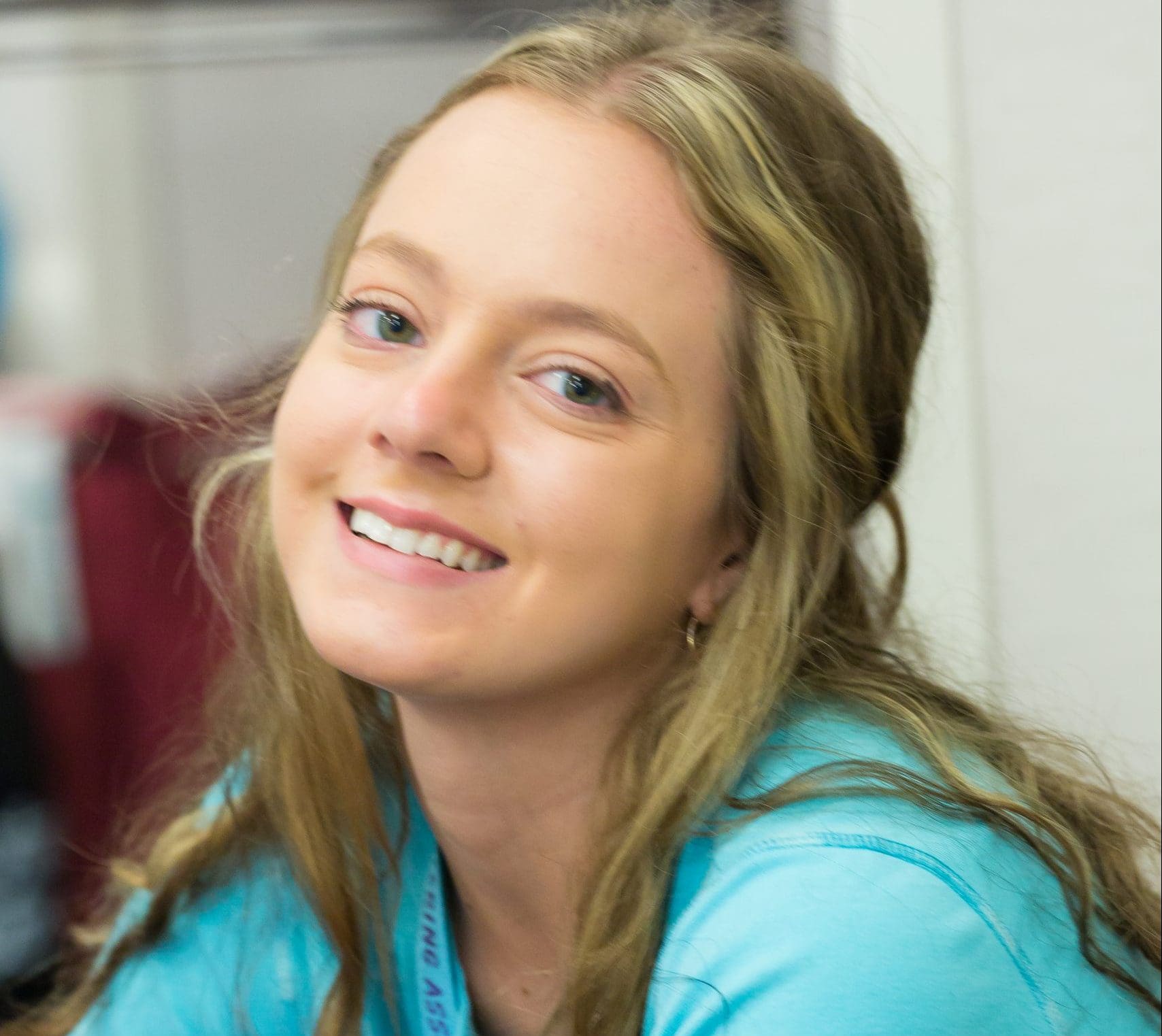 A portrait of a smiling woman with blue eyes and blonde hair, wearing a turquoise "Back to School" shirt, inside a room.