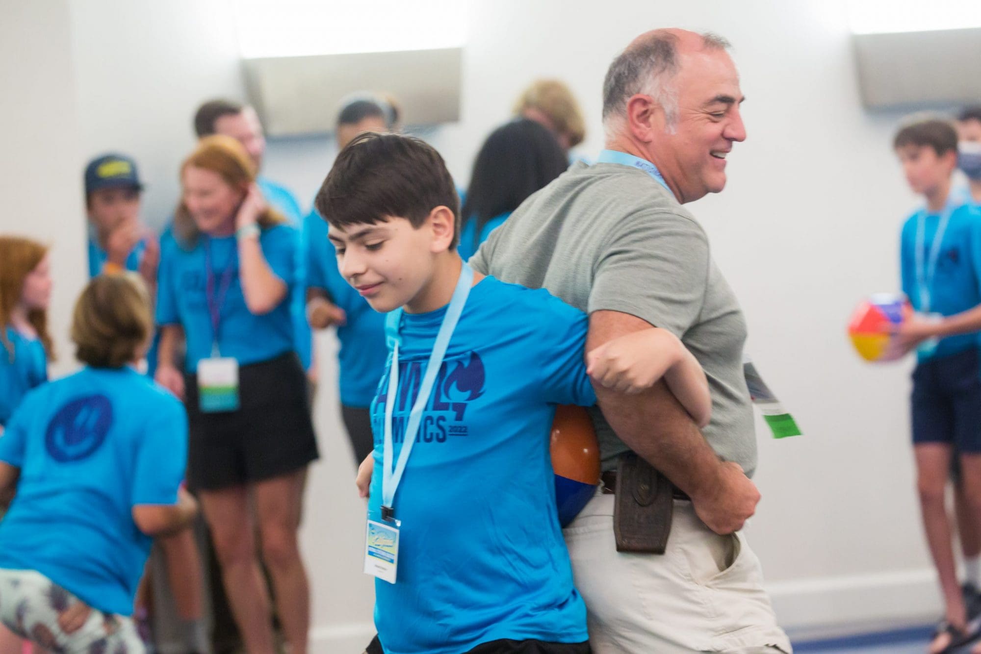 A joyful father embracing his young son at a crowded indoor event. Both are smiling and wearing blue shirts with lanyards, indicating they are part of a group or attending a specific function.