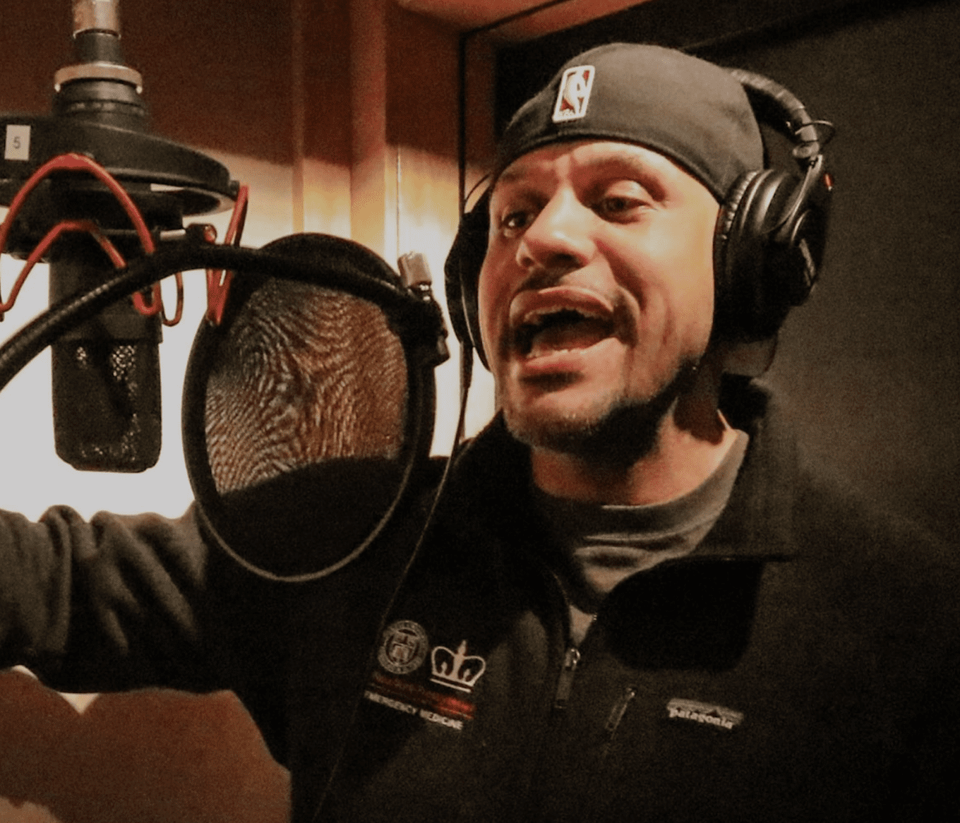 A man wearing a beanie and headphones enthusiastically records a song in a studio, standing in front of a microphone with a pop filter.