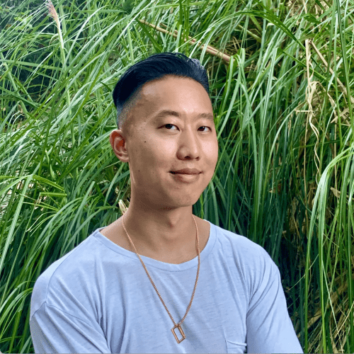 A man with a neat undercut hairstyle smiles gently at a "meet the team" event, wearing a white t-shirt and a gold necklace, positioned against a lush backdrop of green grasses.