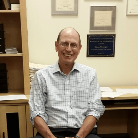 A smiling, balding man wearing glasses and a checkered shirt, sitting in an office with diplomas on the wall behind him, attending the Amplify Conference Portland 2023.