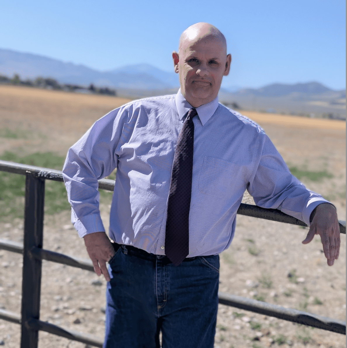 Meet the team: A bald man in a purple shirt and tie stands outdoors, leaning on a railing with mountains in the background. He is wearing jeans and is enjoying the sunlit field.