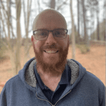 A smiling, bald man with a full red beard, wearing glasses and a blue zip-up jacket, stands outdoors with trees and a cloudy sky in the background. Sean Hare