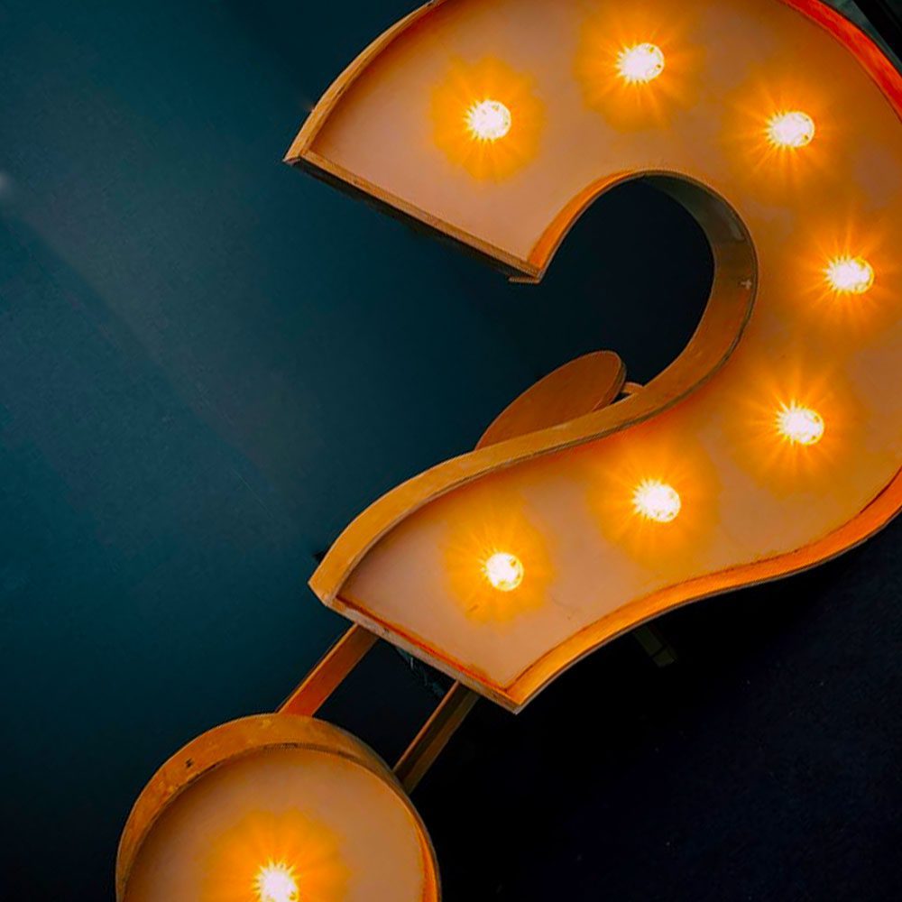 A close-up of a neon-lit question mark sign with glowing orange bulbs against a dark blue background, highlighting the curves and illumination points of the symbol during night tours.
